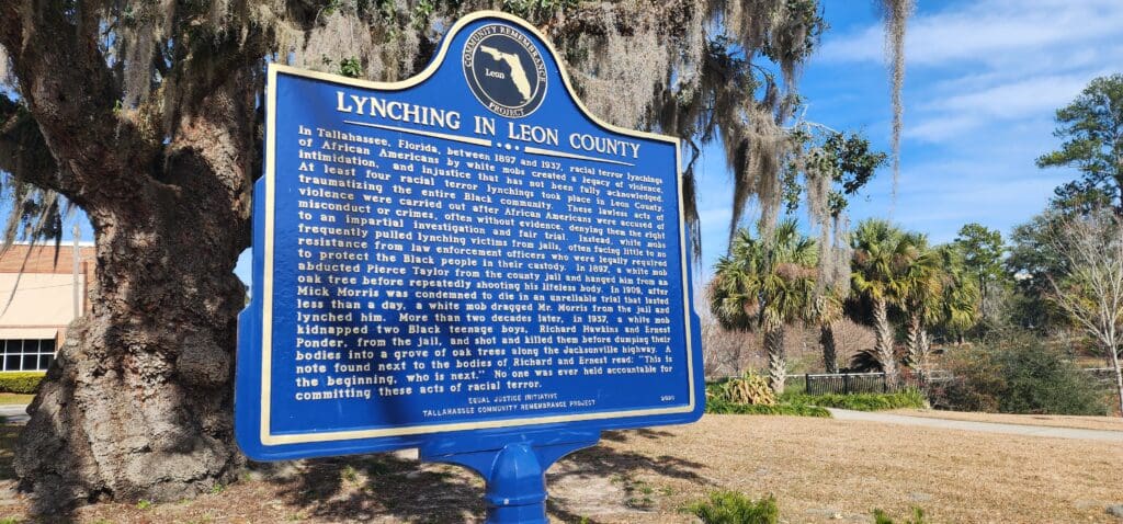 A blue sign in front of trees that has a headline "Lynching in Leon County."