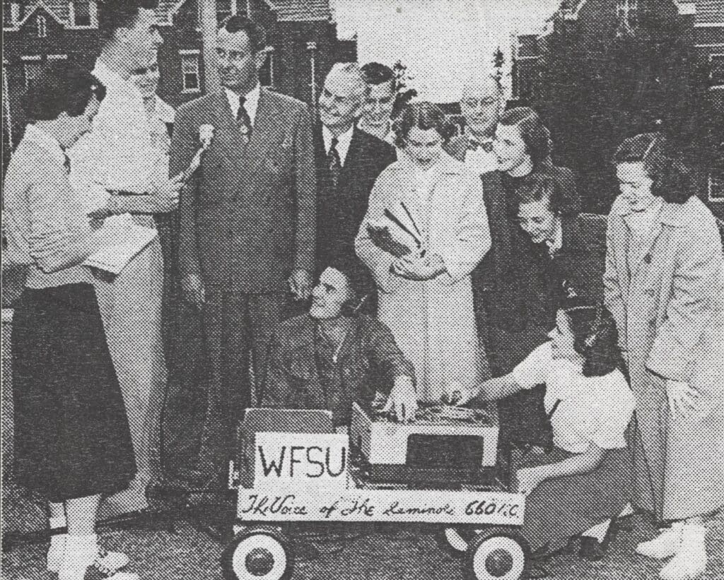 A vintage photo of a group of people around each other with a small wagon with equipment and the words WFSU The Voice of the Seminole 660 AM