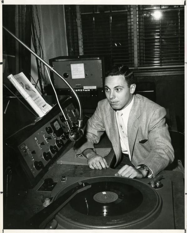 A man sitting in front of a turntable and radio broadcast board.