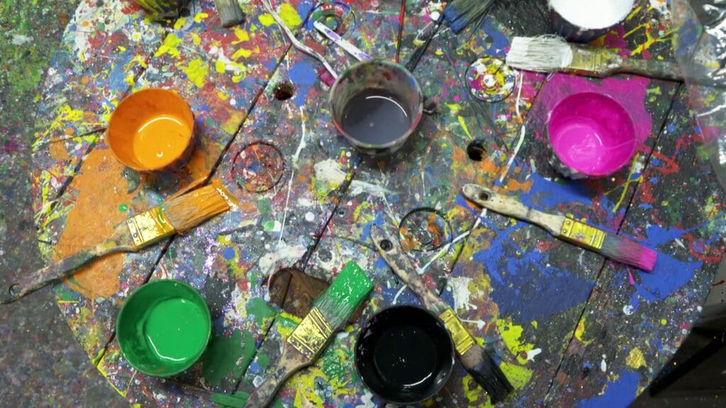 View from above of a wooden table splattered with paint. Paint cups in different colors and brushes sit on the table.