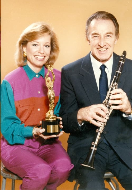 Two people sitting posing for the camera. women on left is holding an award, Man on left is holding a clarinet