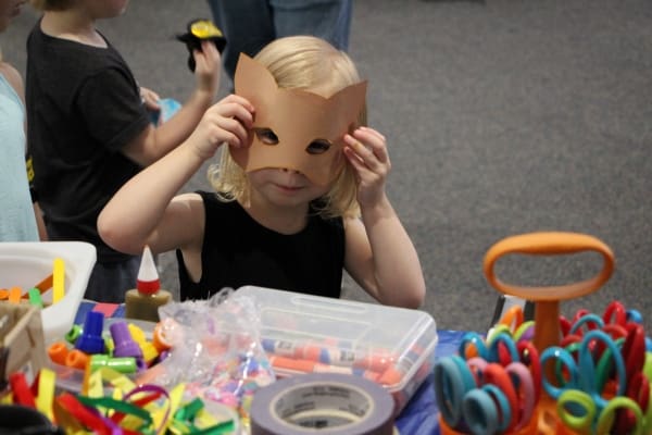 A small child sitting next to table with art equipment around her and construction paper mask on her face.