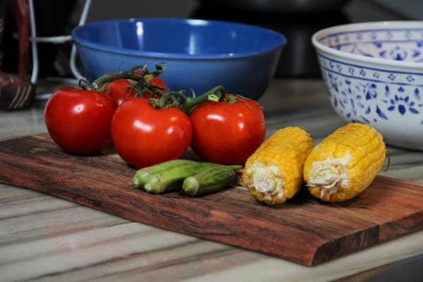 Two tomatoes sitting on top of a wooden cutting board with okra and corn cobs