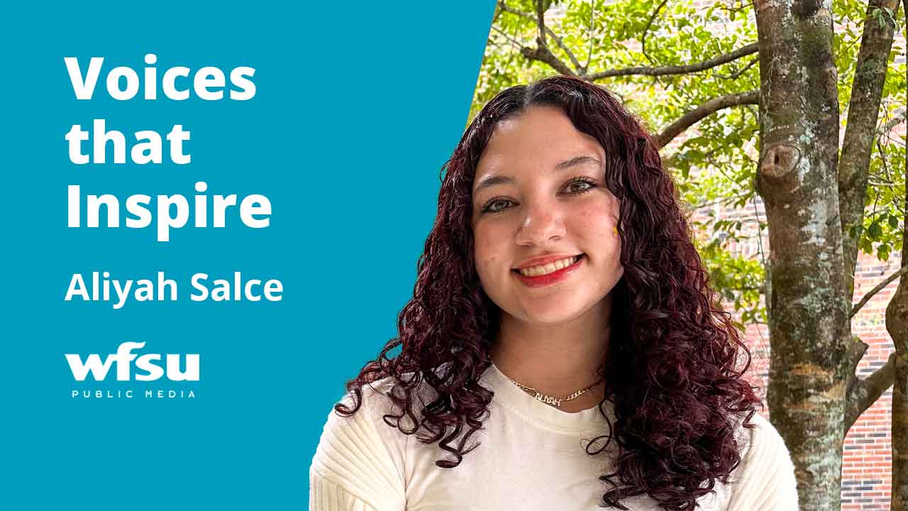 A smiling woman stands in front of a tree and a red brick wall.