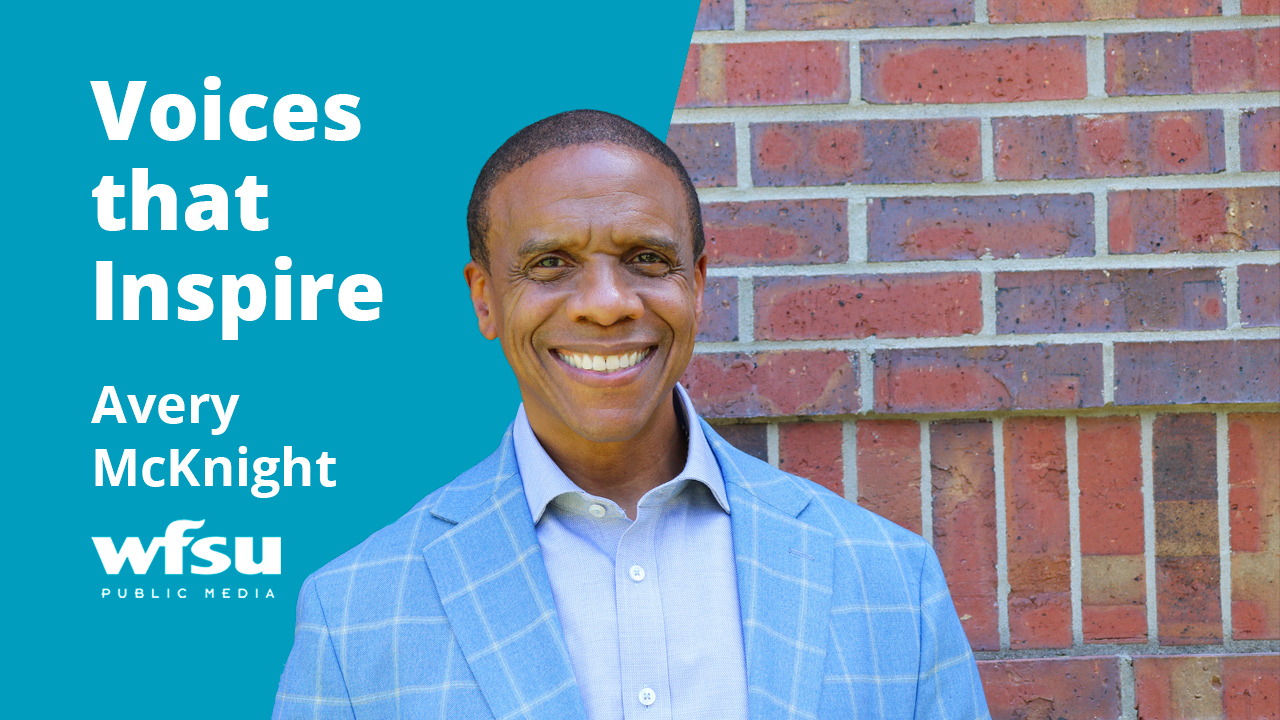 A smiling man in a blue suit stands in front of a red brick wall.