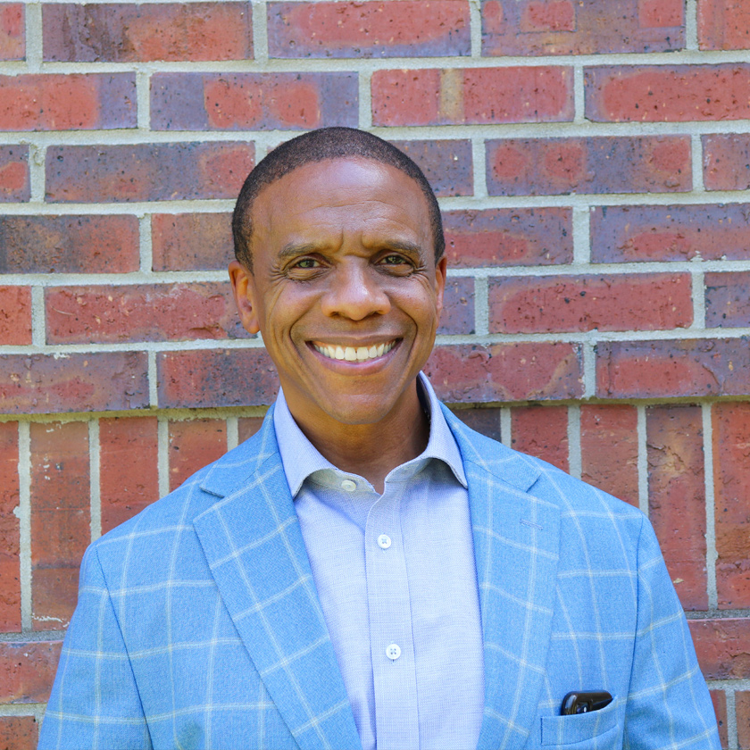 A smiling man in a blue suit stands in front of a red brick wall.