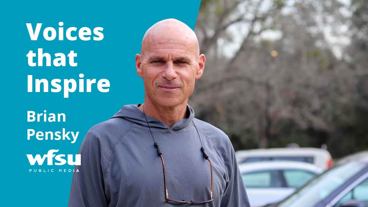 A man in a grey hoodie stands in front of shrubbery.