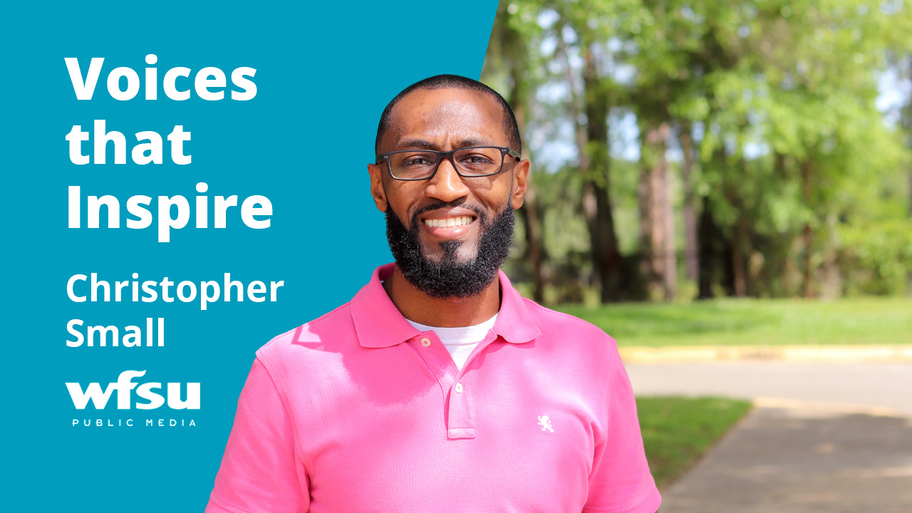 A smiling man in a pink shirt stands outside in front of shrubbery.