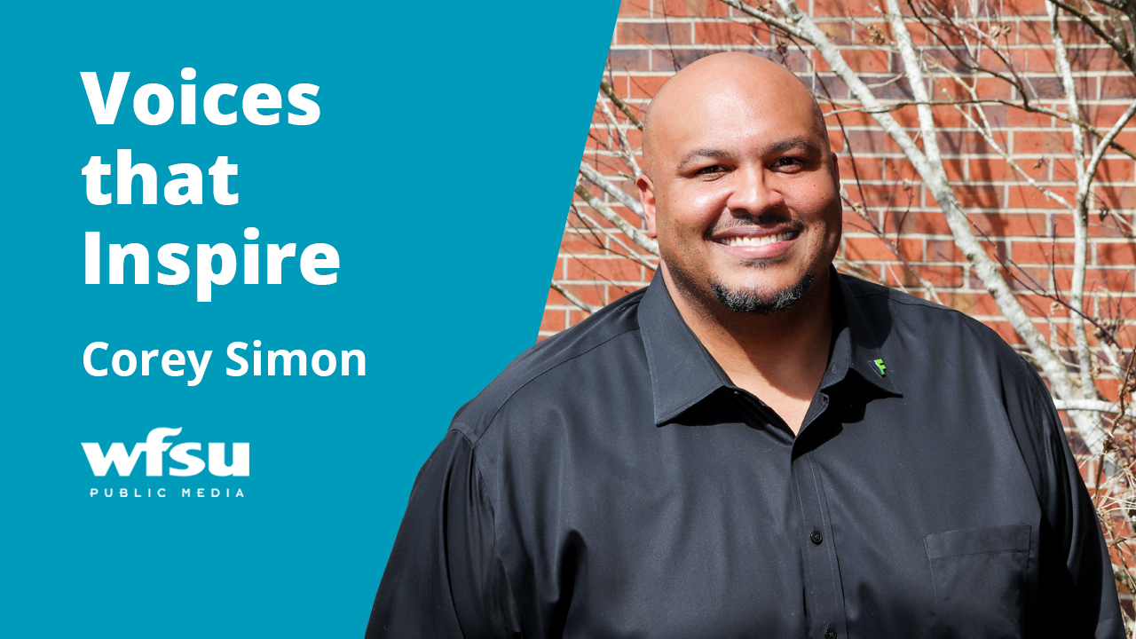 A smiling man in a dark shirt stands in front of a red brick wall.