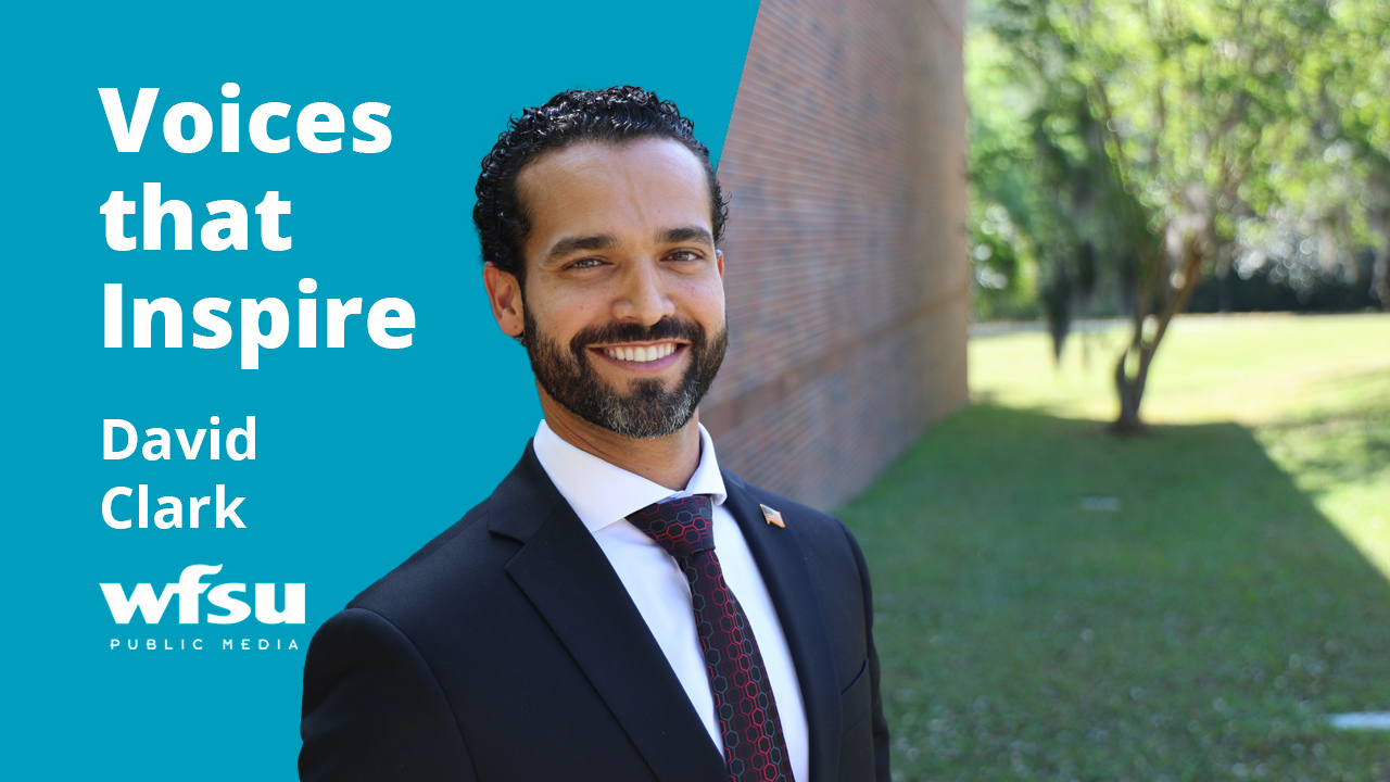 A smiling man in a dark suit stands in front of a red brick wall.