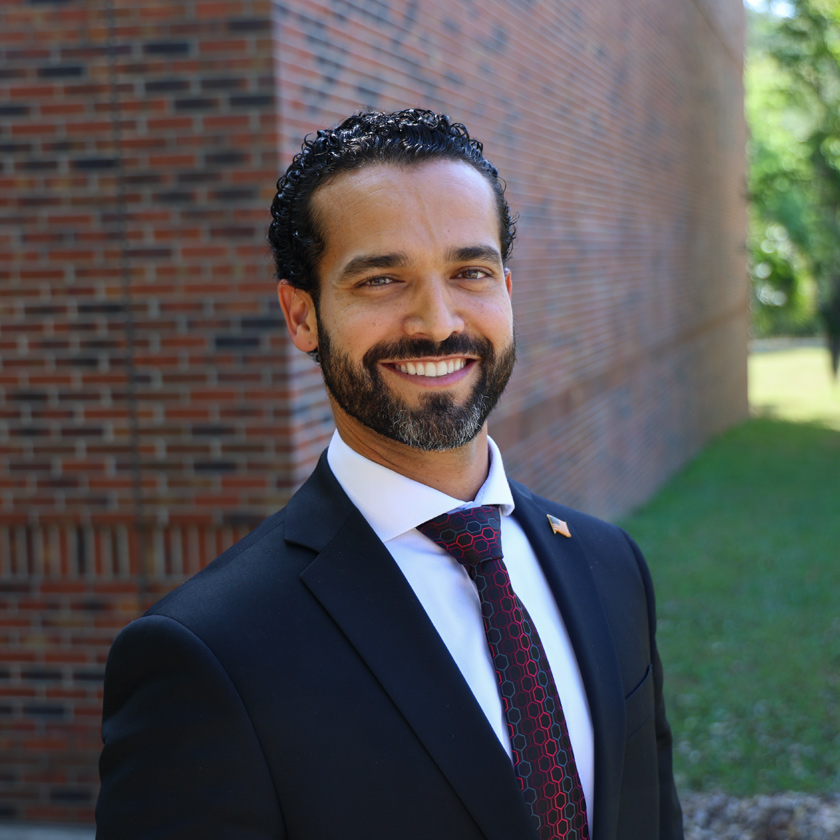 A smiling man in a dark suit stands in front of a red brick wall.