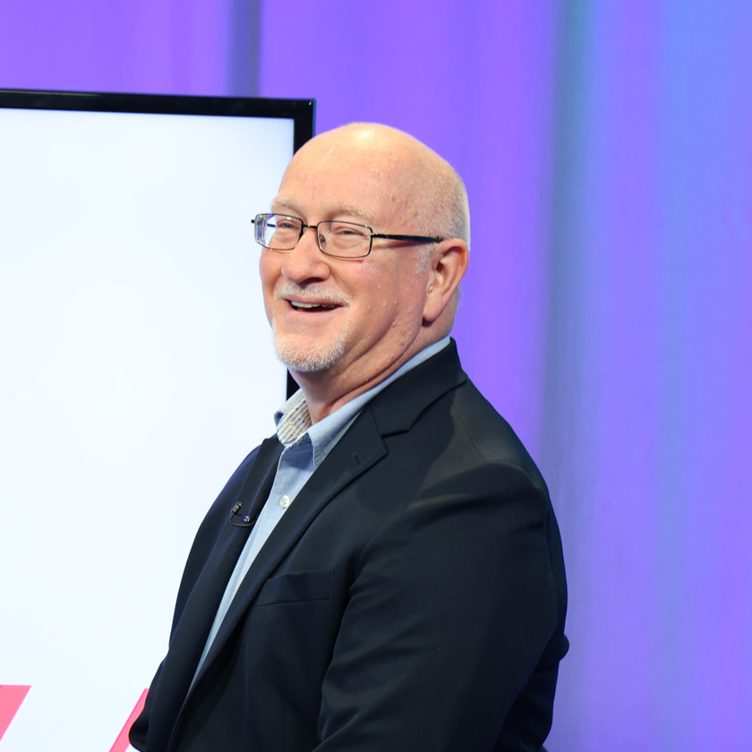 A smiling man in glasses stands in front of a purple screen.