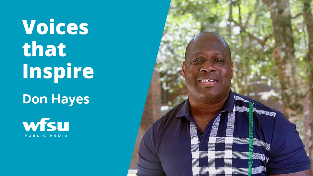 A smiling man in a blue shirt stands in front of a brick wall and a tree.