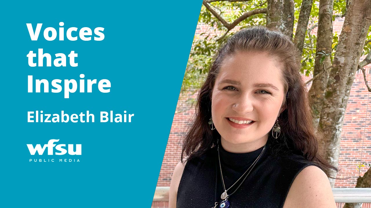 A smiling woman stands in front of shrubbery and a red-brick wall.