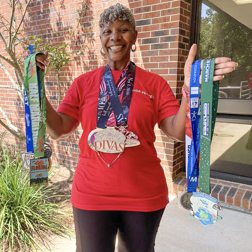 A smiling woman in a red shirt is holding several medals in both of her hands.
