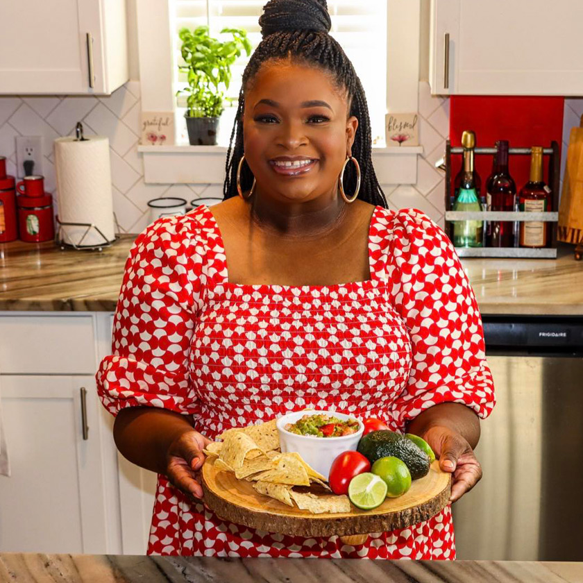 A smiling woman dressed in red and white stands in a kitchen holding a plate of food.