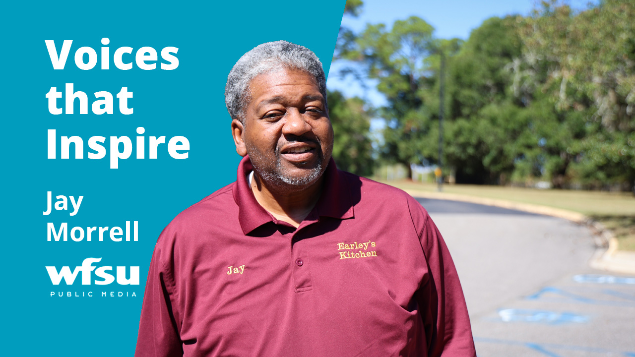 A smiling man in a burgundy shirt stands in front of trees.