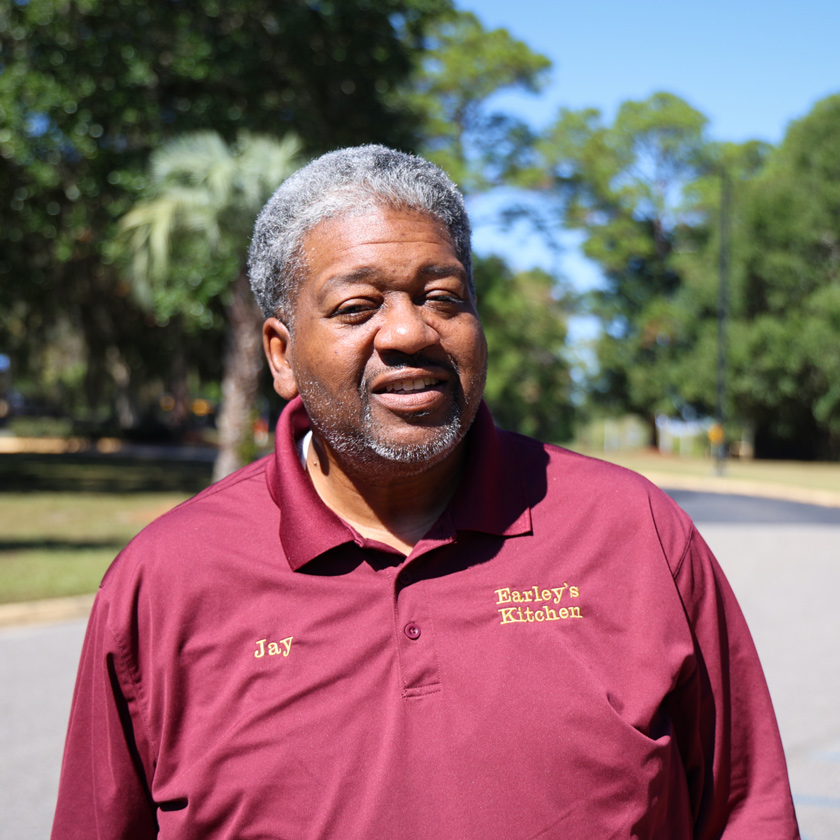 A smiling man in a burgundy shirt stands in front of trees.
