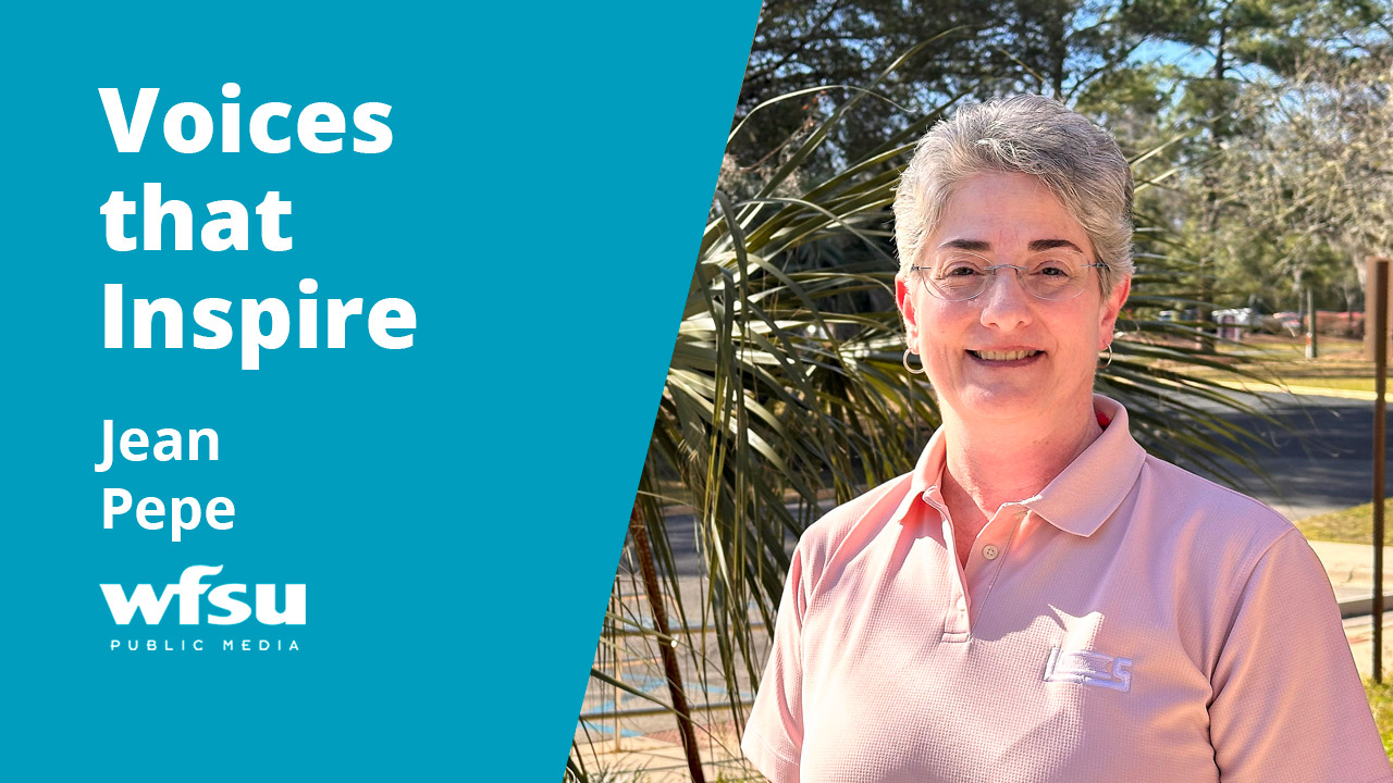 A smiling woman in a pink shirt stands in front of shrubbery.