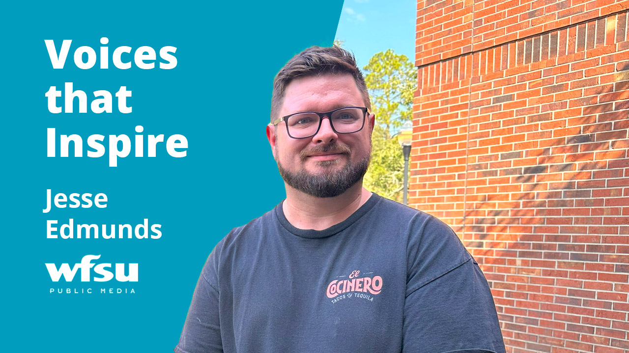 A smiling man stands in front of a brick wall and shrubbery.