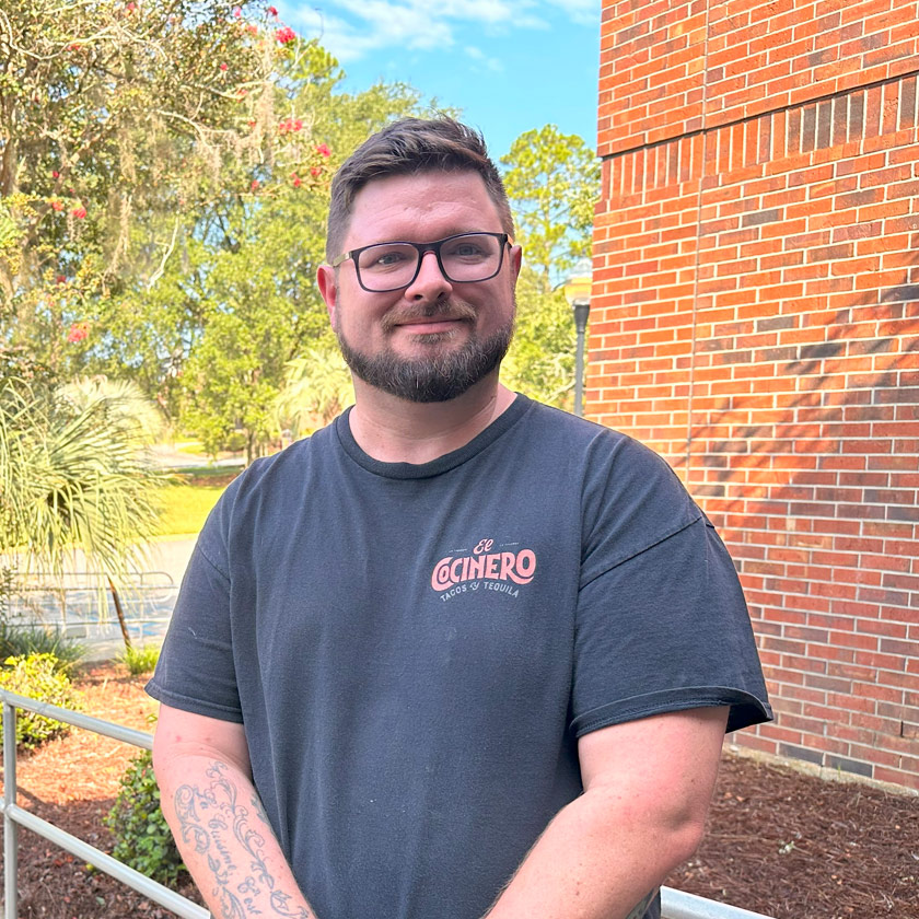 A smiling man stands in front of a brick wall and shrubbery.