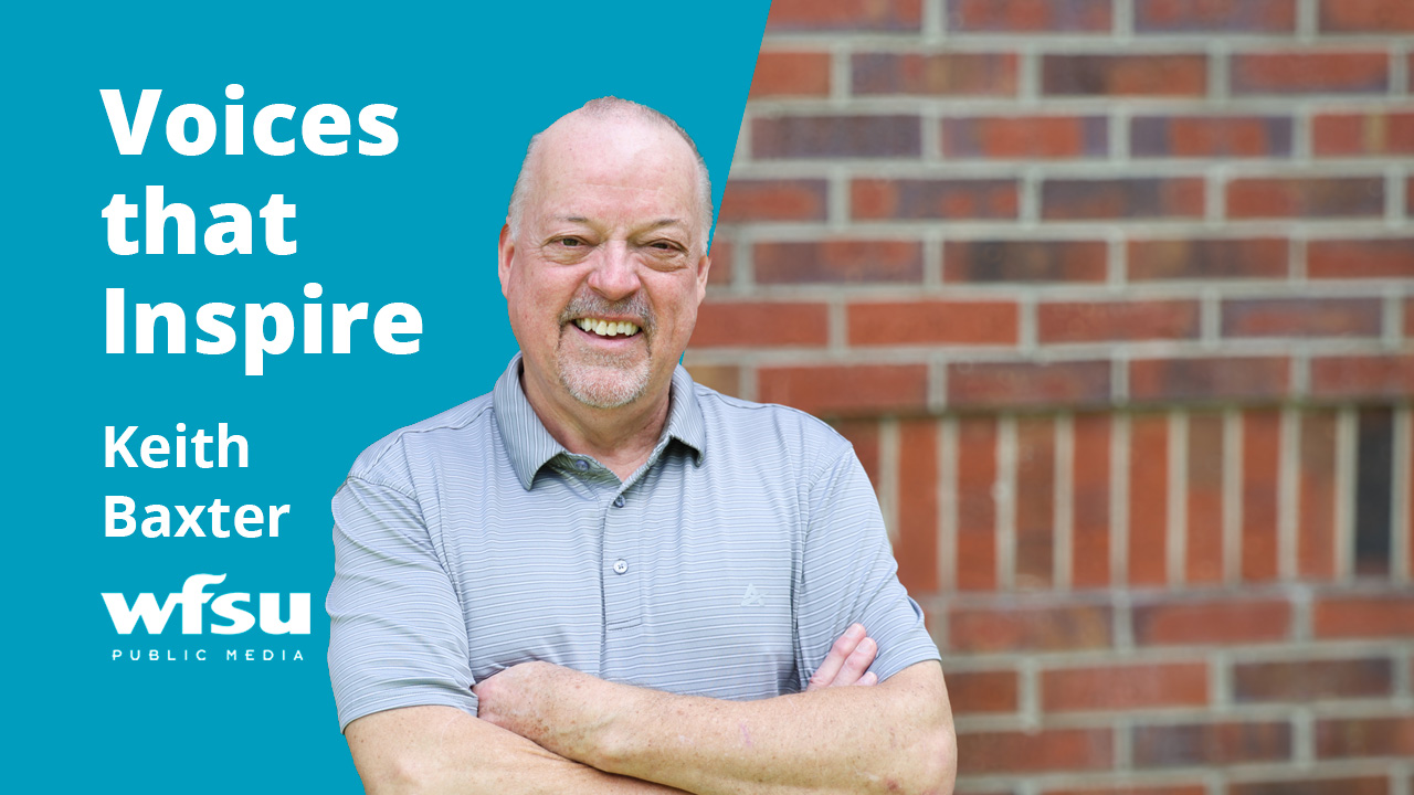 A smiling man stands in front of a red brick wall.