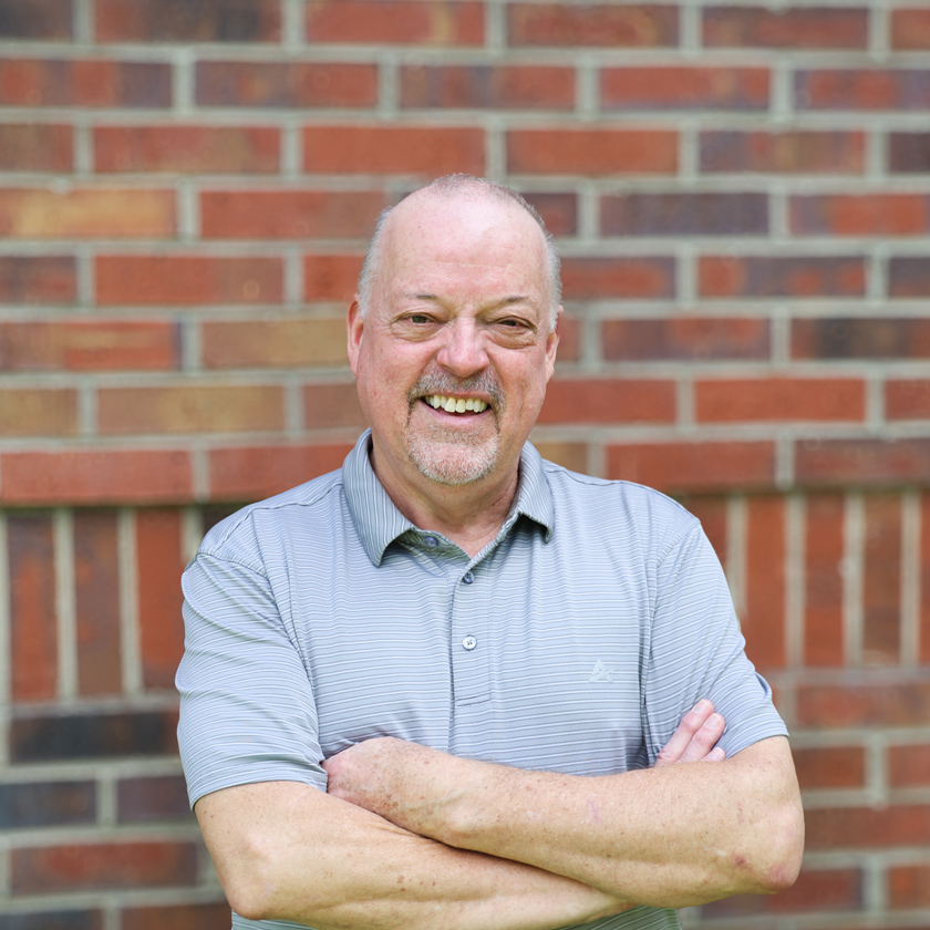 A smiling man stands in front of a red brick wall.