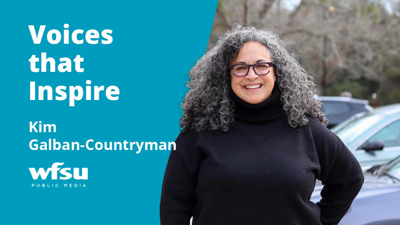 A smiling woman in a black sweater stands outside in front of shrubbery.