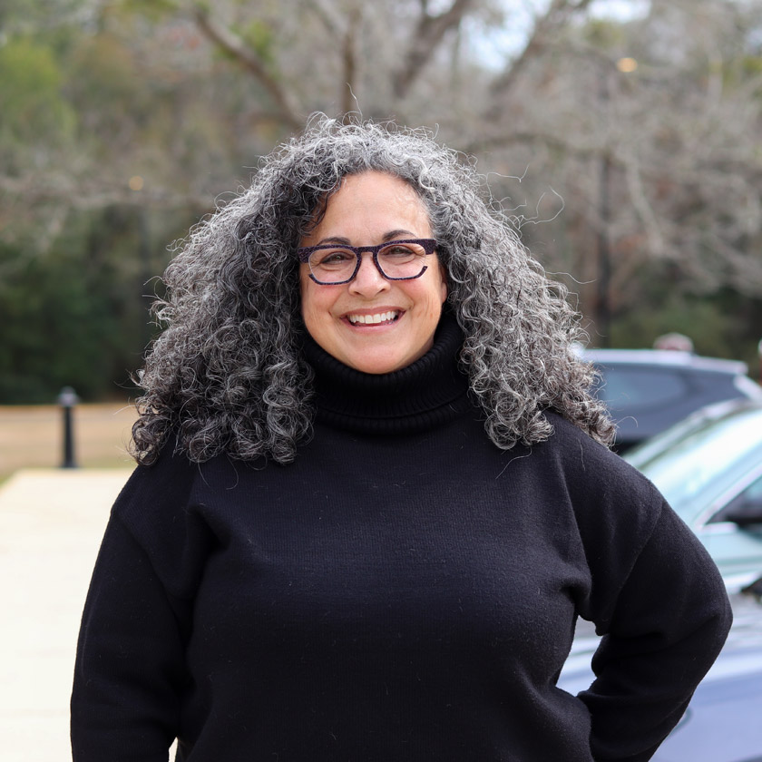 A smiling woman in a black sweater stands outside in front of shrubbery.