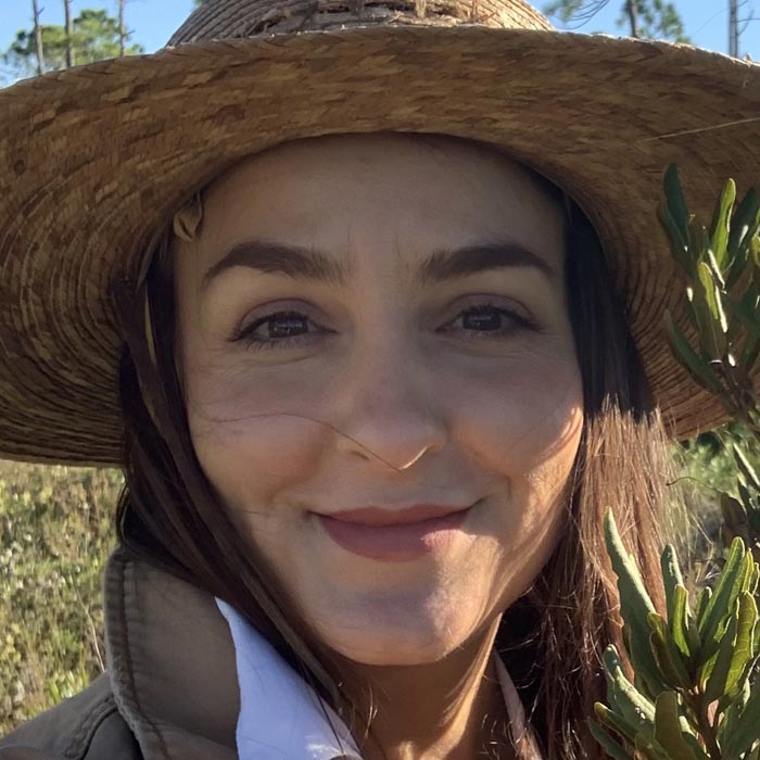 A smiling woman standing in nature wearing a sun hat 