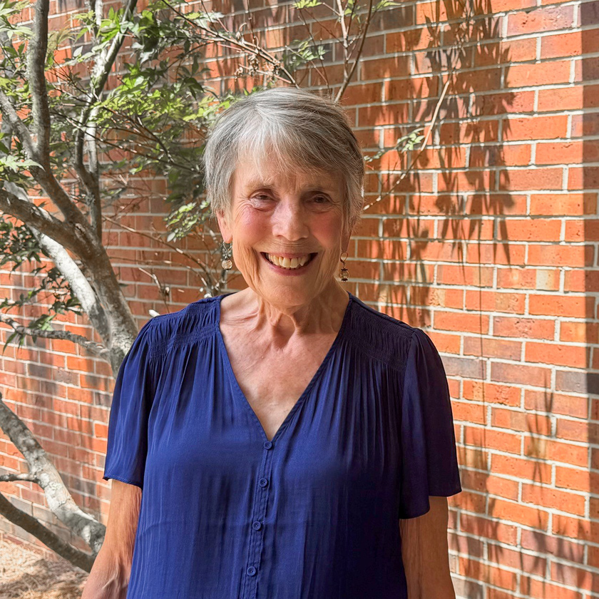 A smiling woman in a blue shirt stands in front of a red-brick wall.