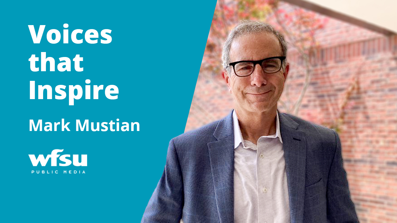 A smiling man in a blue suit stands outside in front of a red brick wall