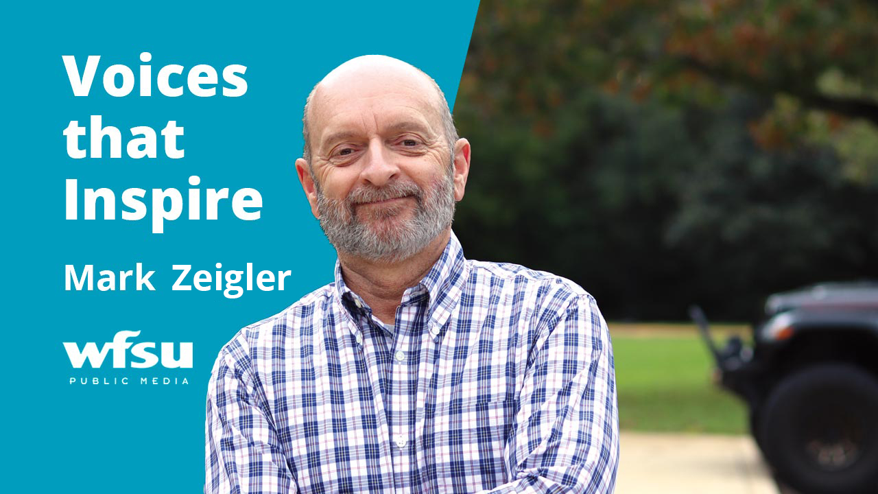 A smiling man in a blue and white checkered shirt smiles.