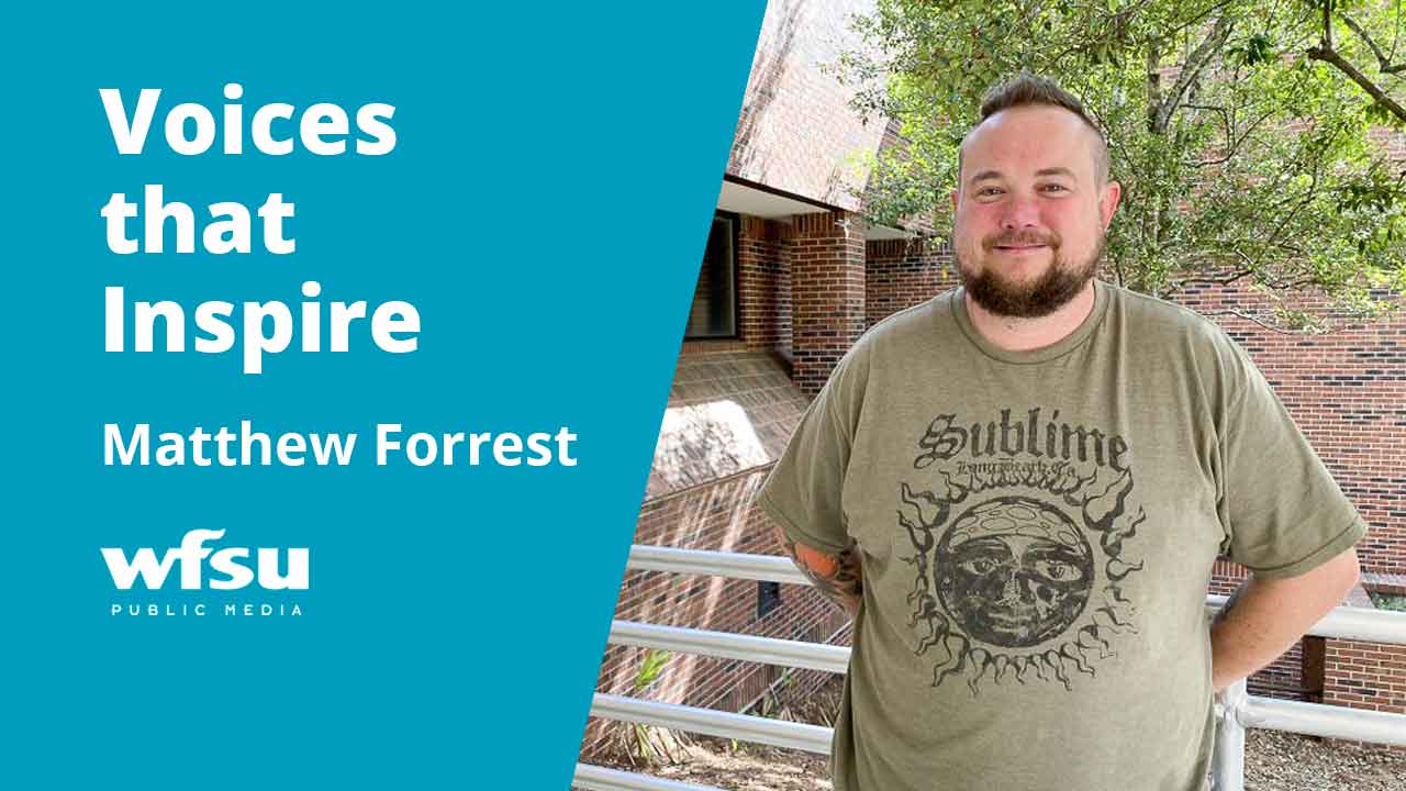 A smiling man stands in front of shrubbery.