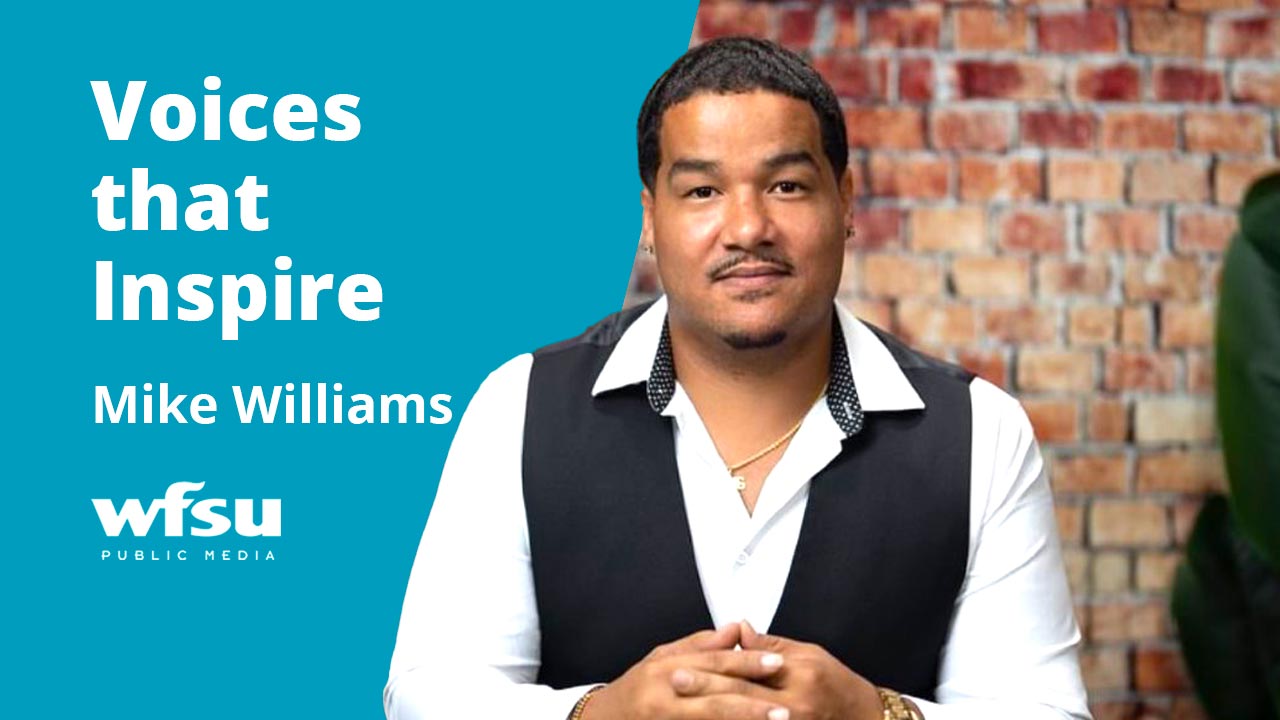 A smiling man sits with hands folded in front of a red brick wall.