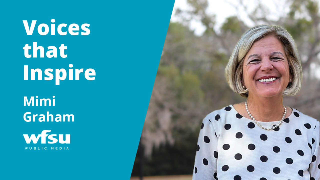 A smiling woman in a white and black top stands in front of shrubbery.
