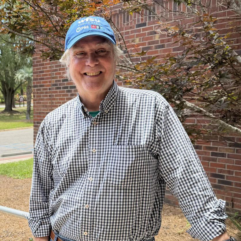 A smiling man in a blue baseball cap stands in front of a red brick wall.