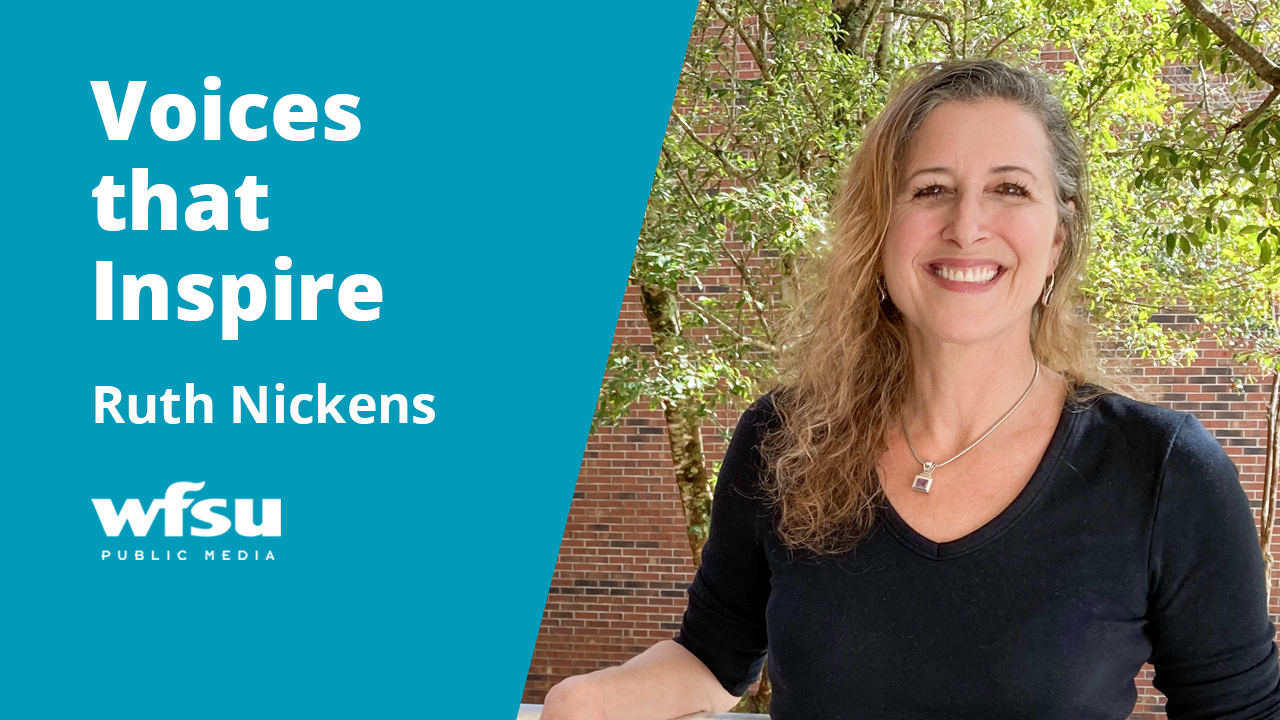 A smiling woman stands in front of a red-brick wall and a tree.