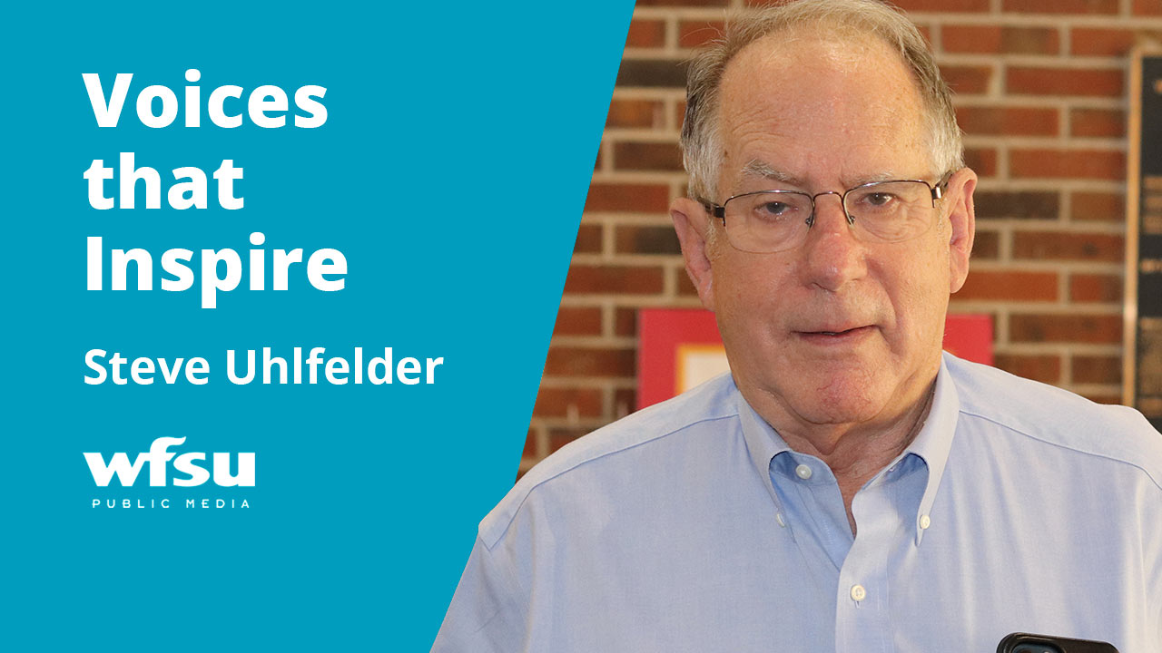 Bespectacled man in light button-up shirt stand in front of a red brick wall.