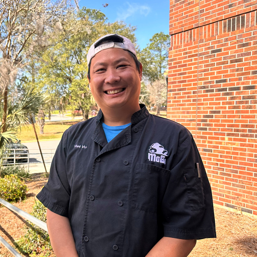 A man smiling stands outside smiling in a black chef's shirt.