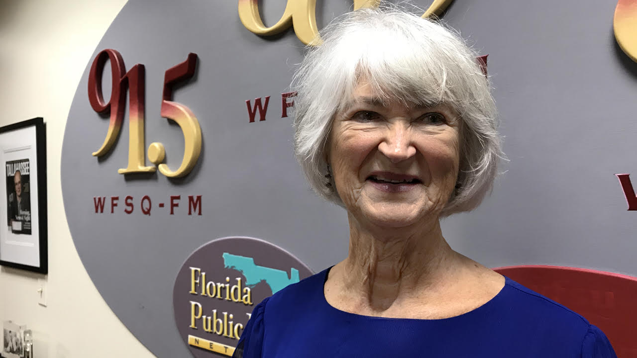 A smiling older, white haired woman in a blue blouse in front of a light purple wall. 