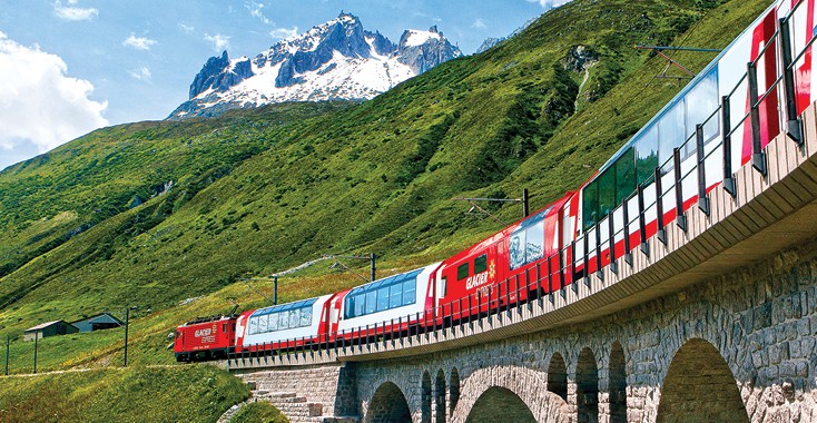 Red train crossing a cobblestone bridge in steep green hills with high mountains seen in the background.