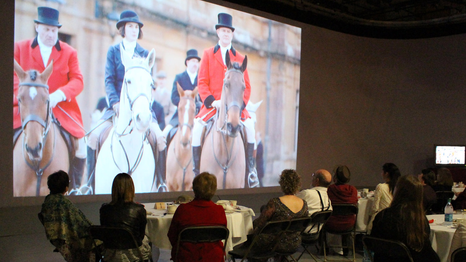 people sitting at round tables watching a large projection of actors on horses