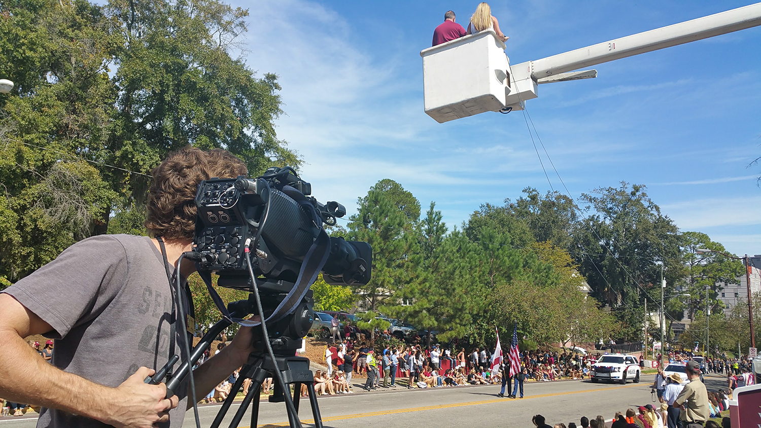a male student looking through the viewfinder of a video camera on a street where a parade is about to take place