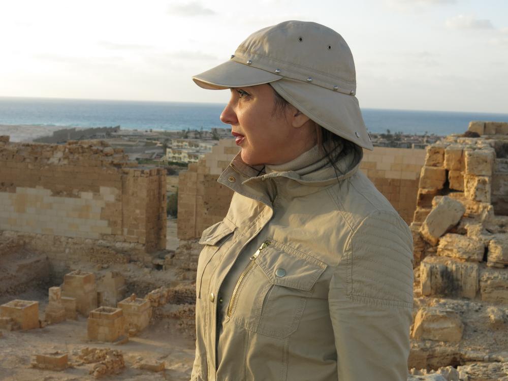Picture of an archaeologist standing in some ancient ruins near a giant body of water.