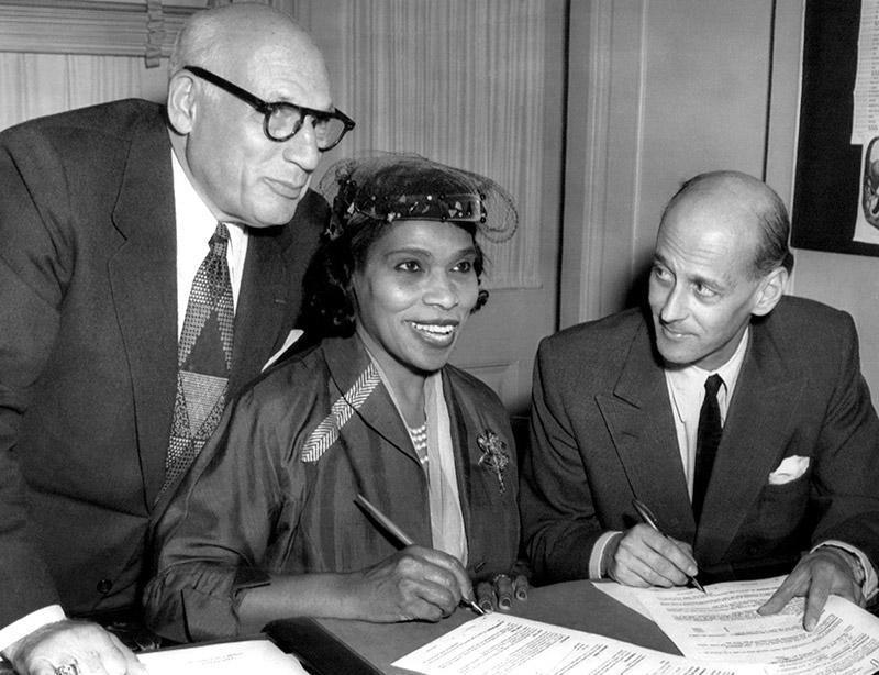 black woman signs document at table - flanked by two white men in suits, mid 20th century dress