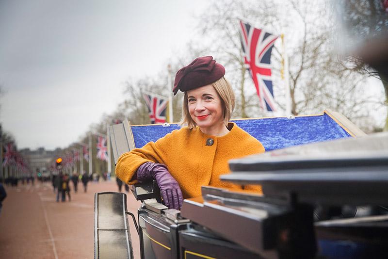 blonde woman in mustard coat sitting in stage coach with British flags hanging behind her. Looks like road leading up to Buckingham palace 